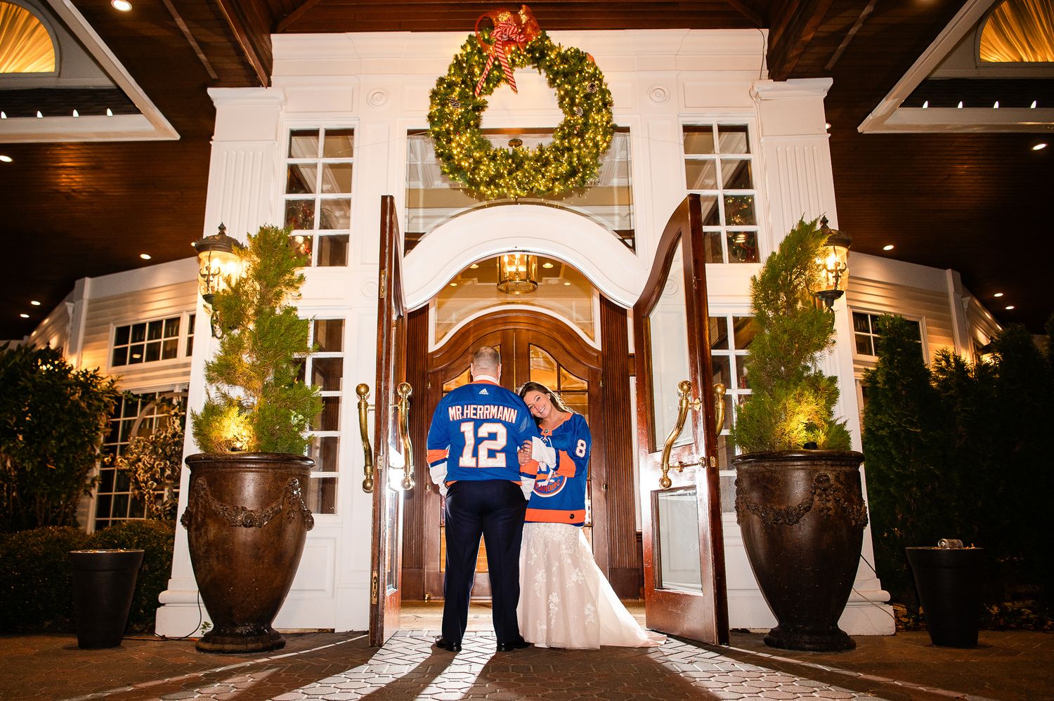A couple in blue sports jerseys stands in front of an elegant white mansion entrance with holiday decor at night.