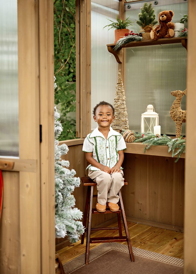 A smiling young boy sits on a stool inside a festively decorated greenhouse with Christmas trees and holiday decor.