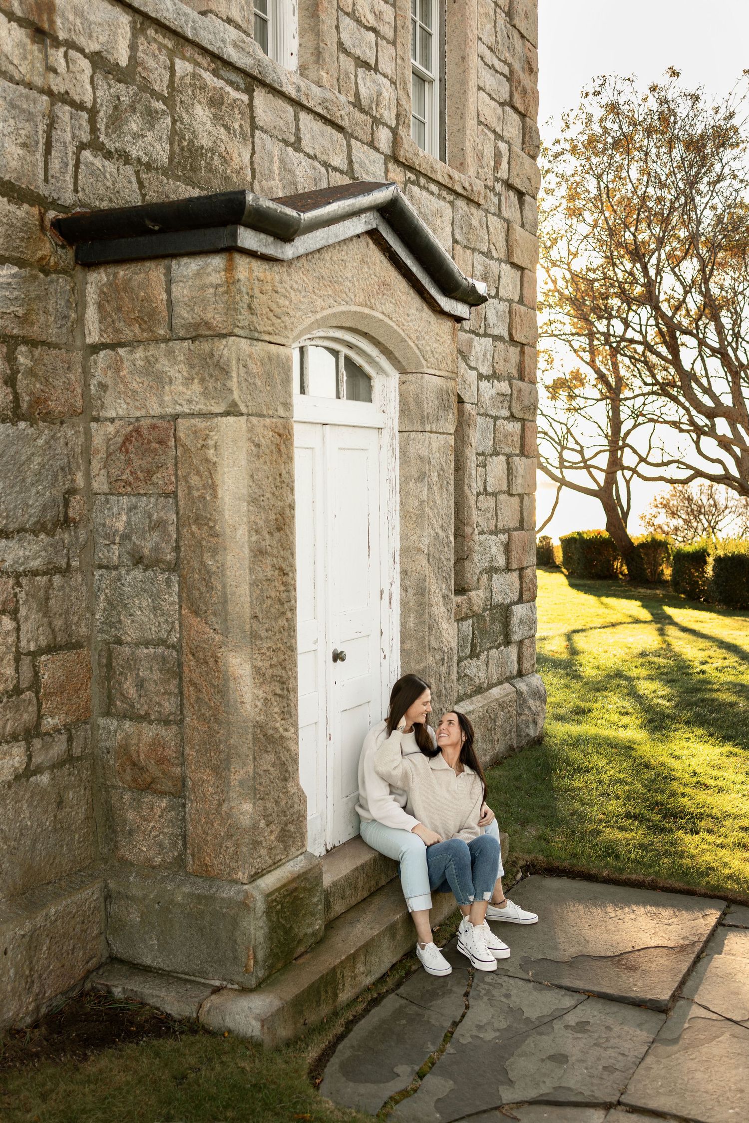 Two women sit close together on the stone steps of a historic granite building with a white door, framed by golden autumn light and trees.