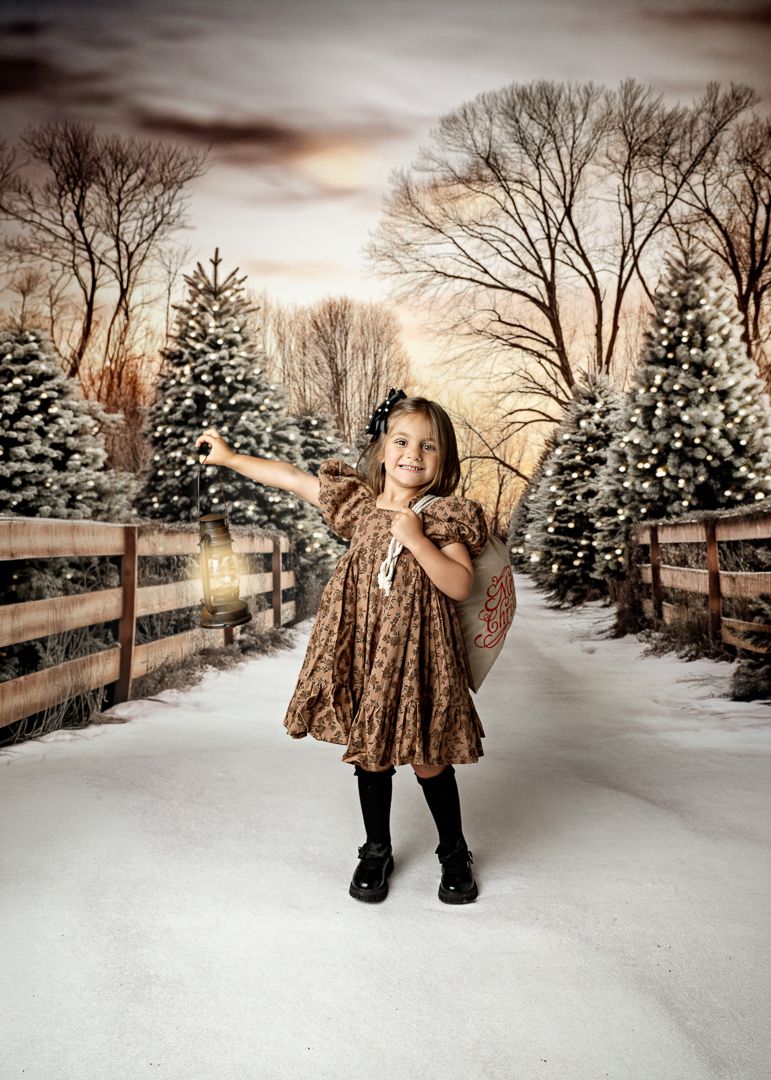 Young girl in floral dress poses gracefully on snowy path lined with Christmas trees and wooden fence.