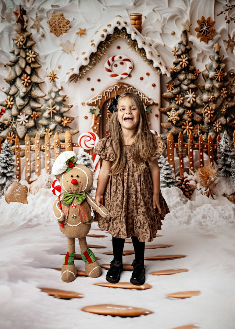 A laughing girl in a brown dress poses with a gingerbread man plush in a festive holiday gingerbread house scene.