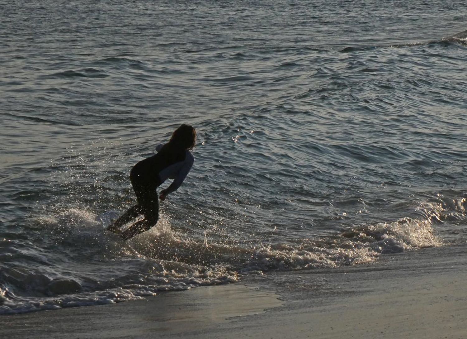 A person skimboarding along the shoreline at sunset, creating splashes in the ocean waves.