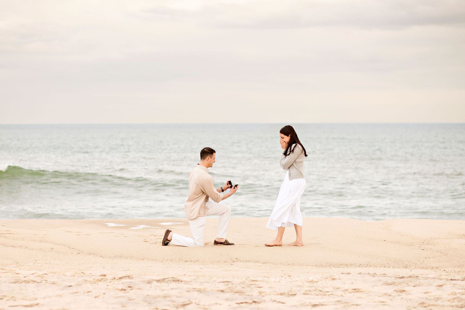 A romantic beach proposal scene with a couple standing on golden sand by the ocean at sunset.