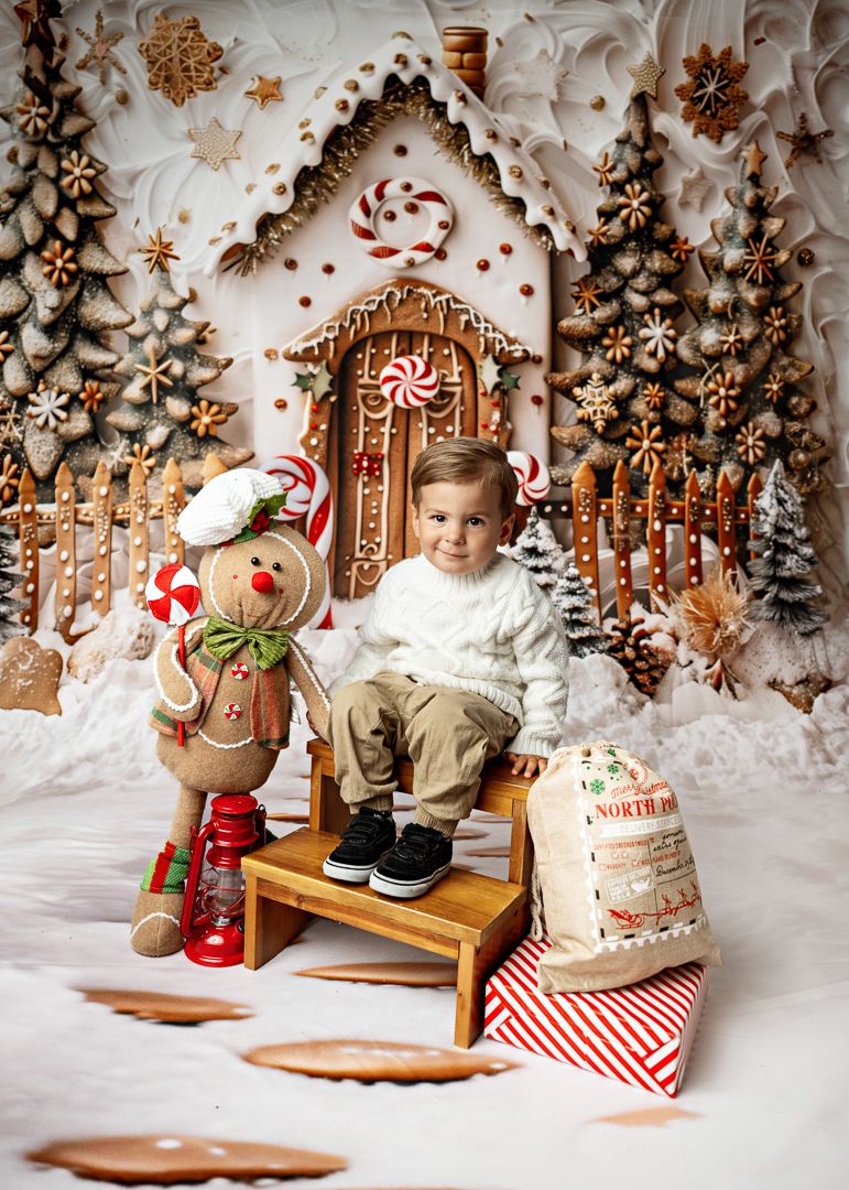 Toddler boy in white sweater sits on bench before gingerbread house backdrop with Santa sack and gingerbread man.