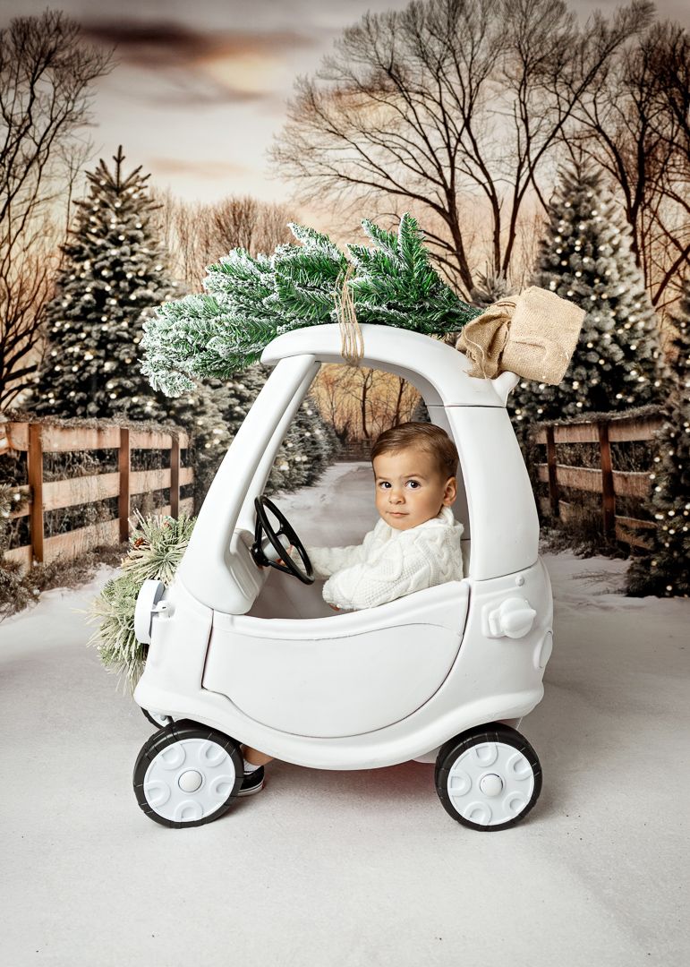 Baby in white cozy coupe toy car with Christmas tree on top, snowy winter holiday photo session backdrop.