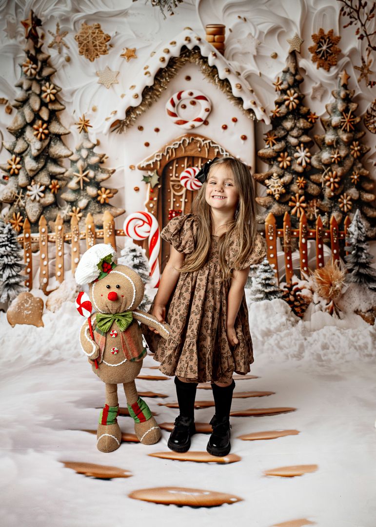 Young girl in brown dress poses with gingerbread man toy before festive candy house Christmas backdrop.