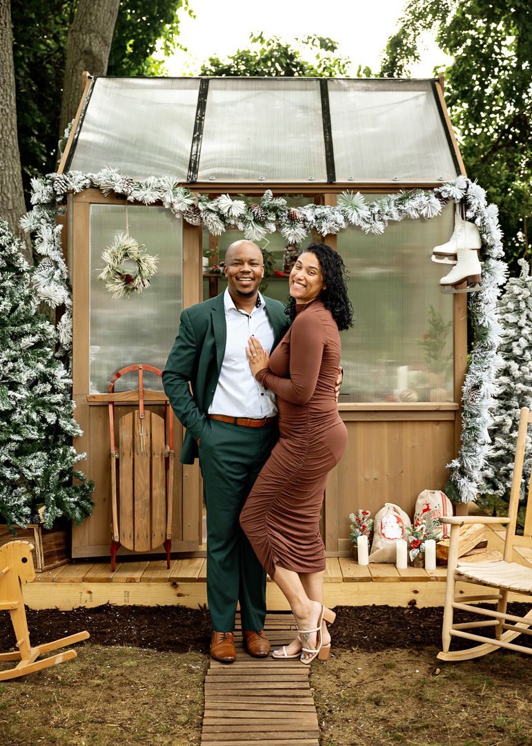 Couple posing in front of a Christmas-decorated greenhouse with holiday ornaments and white trees.
