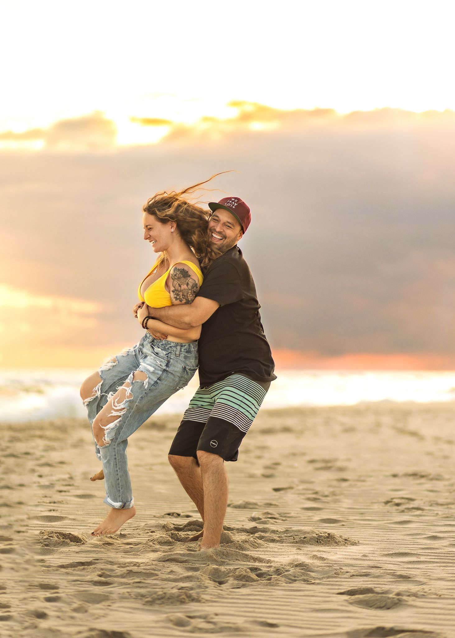 A couple embraces playfully on a sandy beach at sunset with golden light illuminating the romantic scene.