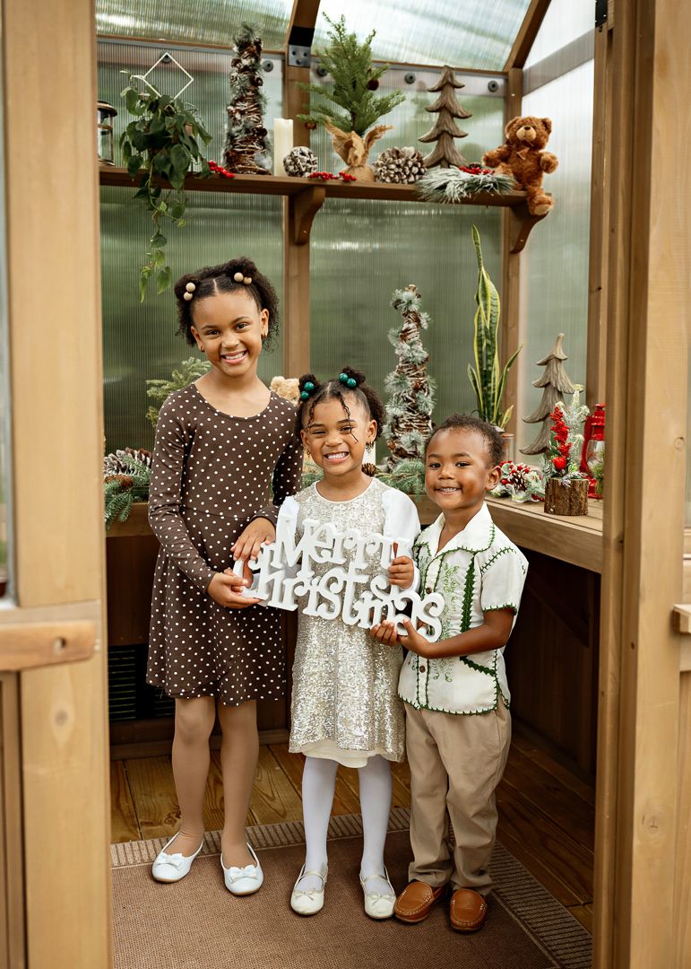 Three children pose with a Merry Christmas sign inside a festive greenhouse decorated with holiday trees and ornaments.