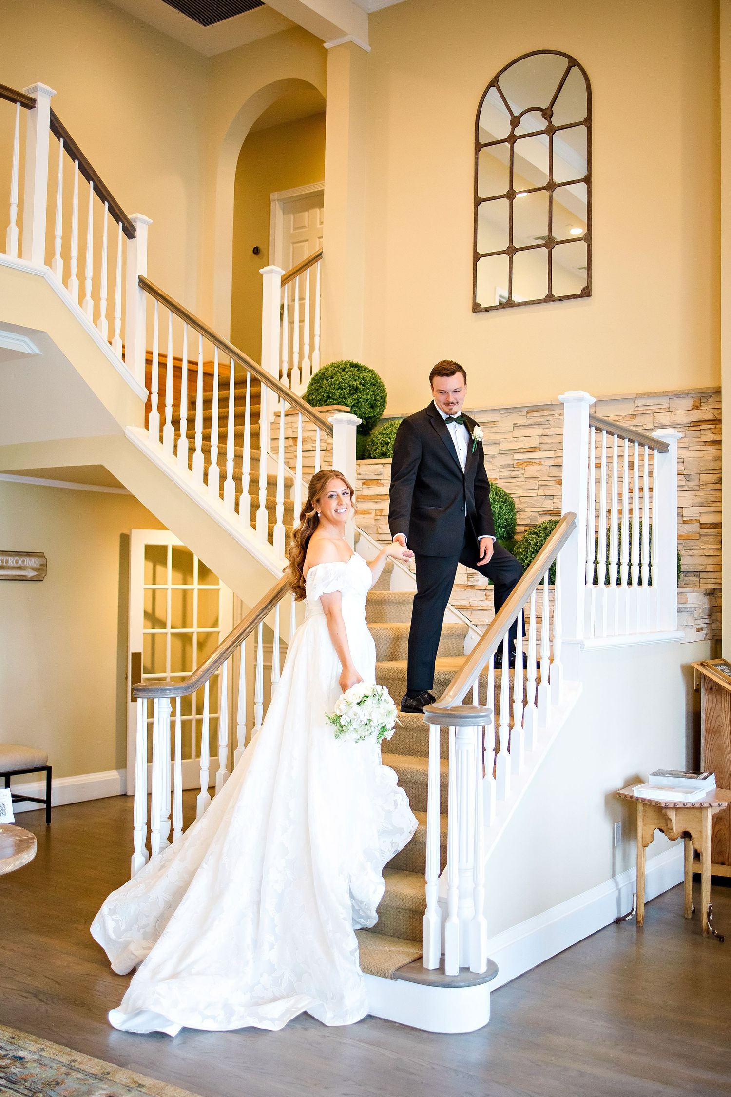 A wedding scene with a bride in white gown and groom in tuxedo posing elegantly on a grand staircase in an interior setting.