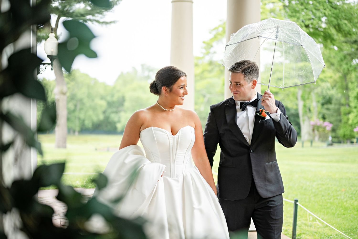 Bride in strapless gown and groom in tuxedo walk under clear umbrella in lush green park on rainy wedding day.
