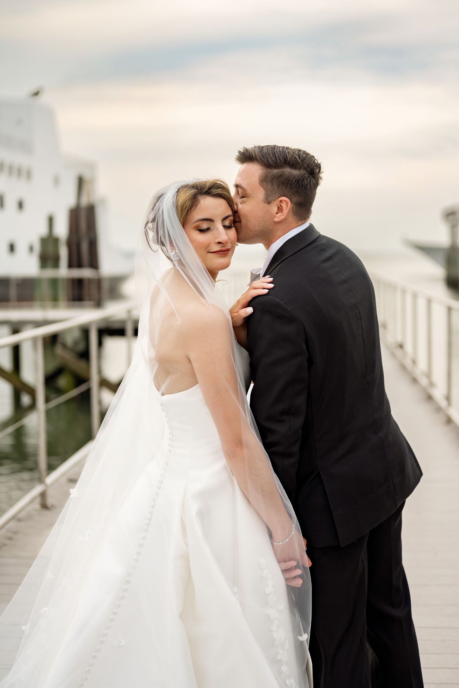 Bride and groom share a romantic kiss on a waterfront pier, bride in white gown with veil, groom in black suit.