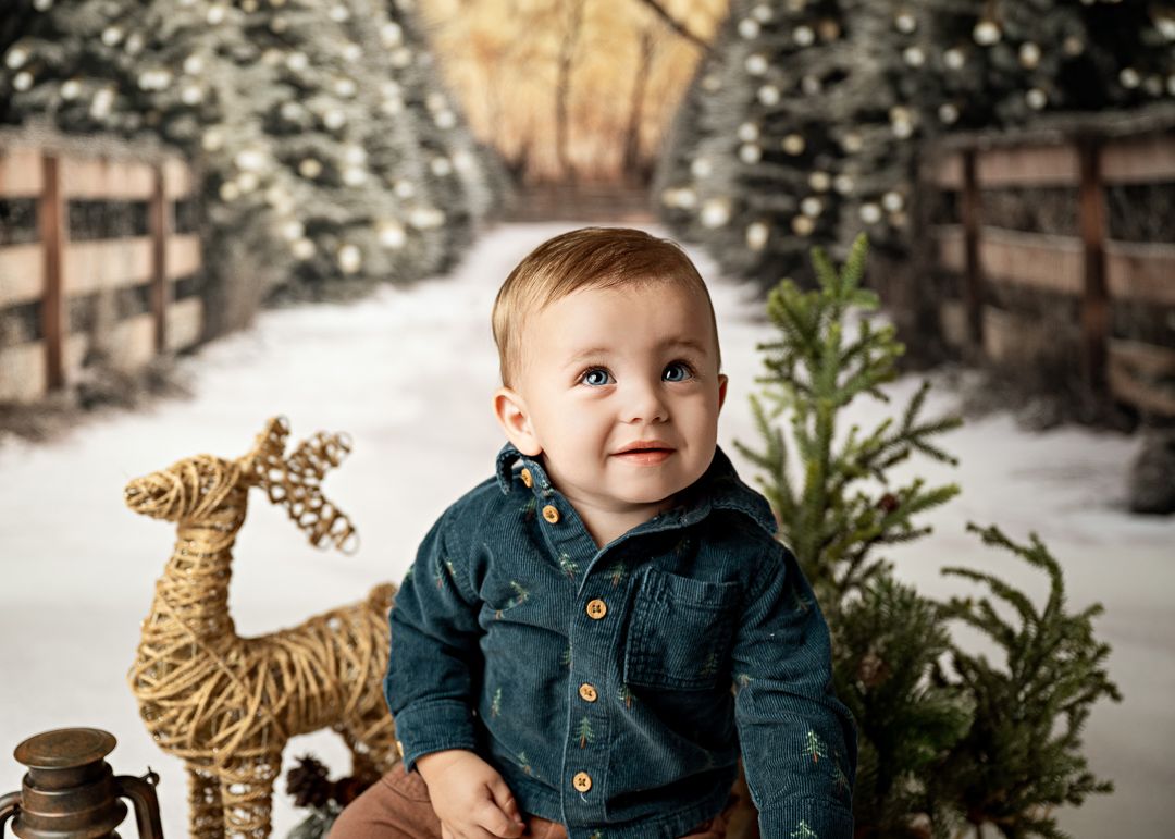 Adorable baby boy in teal shirt poses with reindeer decor and Christmas trees in snowy winter scene.