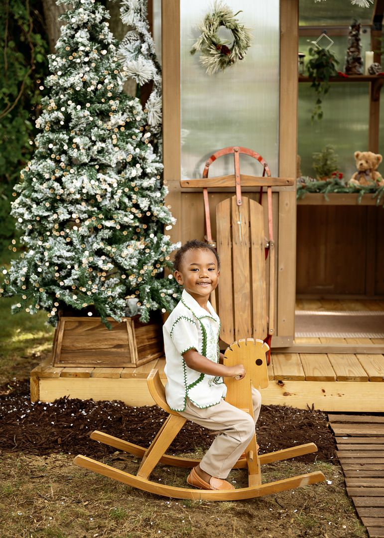 Young child smiles while sitting on a wooden rocking horse beside a snow-flocked Christmas tree.
