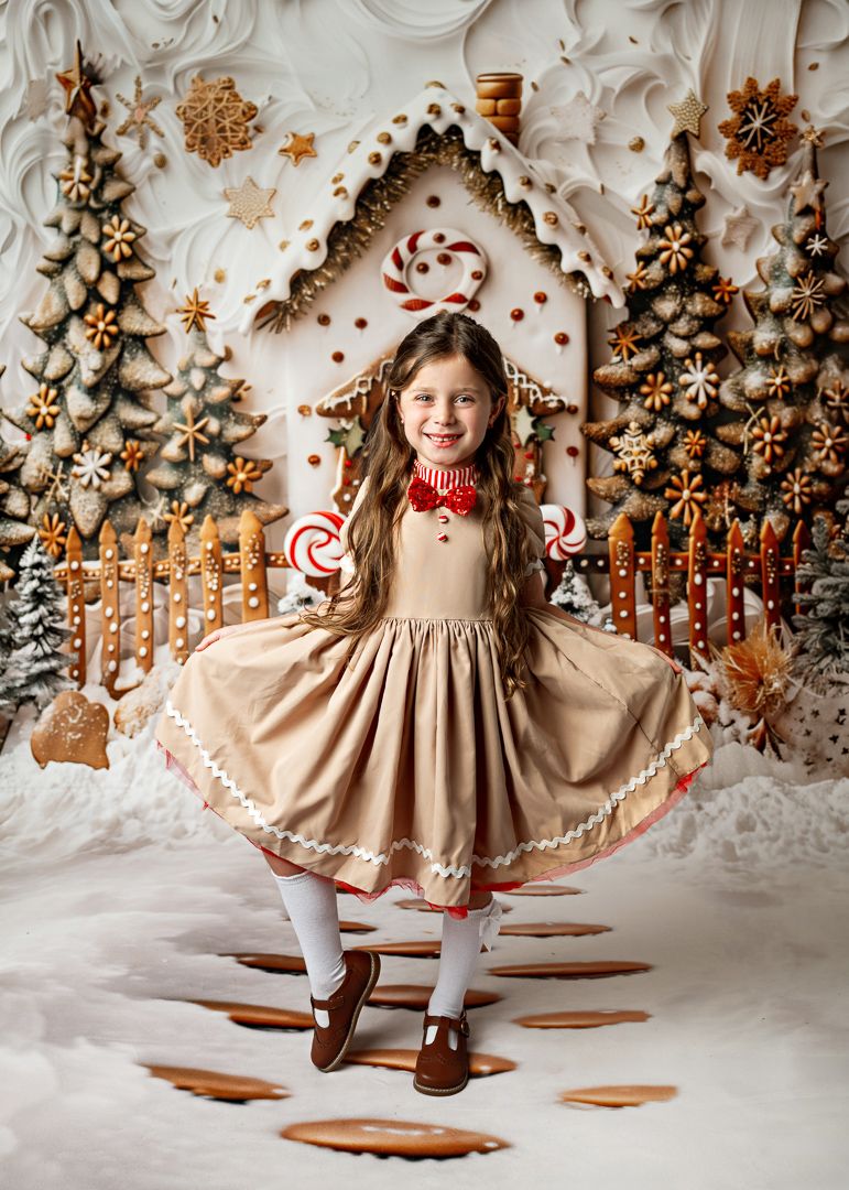 Girl in tan dress poses before a festive gingerbread house backdrop with candy canes and Christmas trees.
