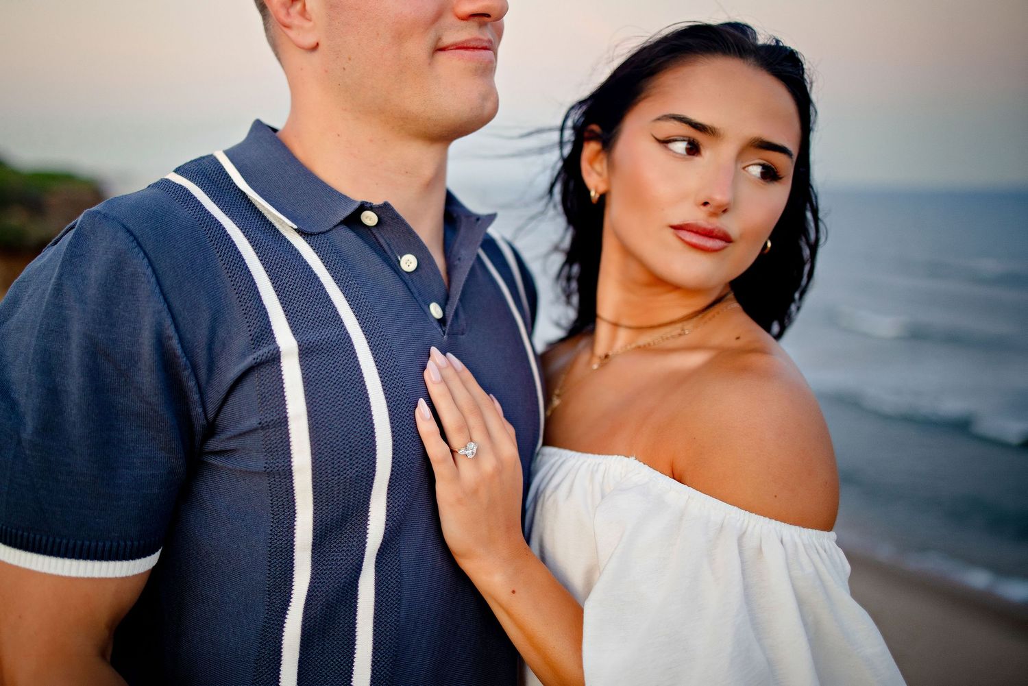 Couple embracing on a beach at sunset, woman in white off-shoulder top with engagement ring, man in blue polo shirt.