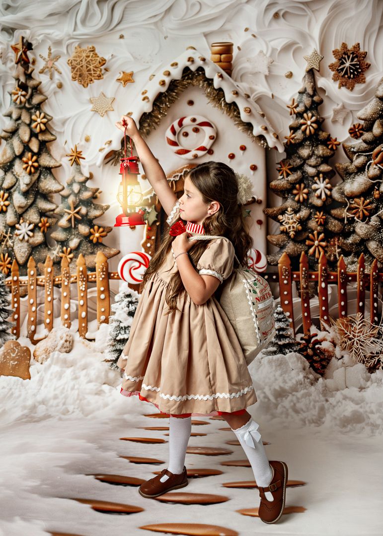 Young girl in a tan dress reaches up to touch a candy cane decoration in an enchanting gingerbread Christmas backdrop.