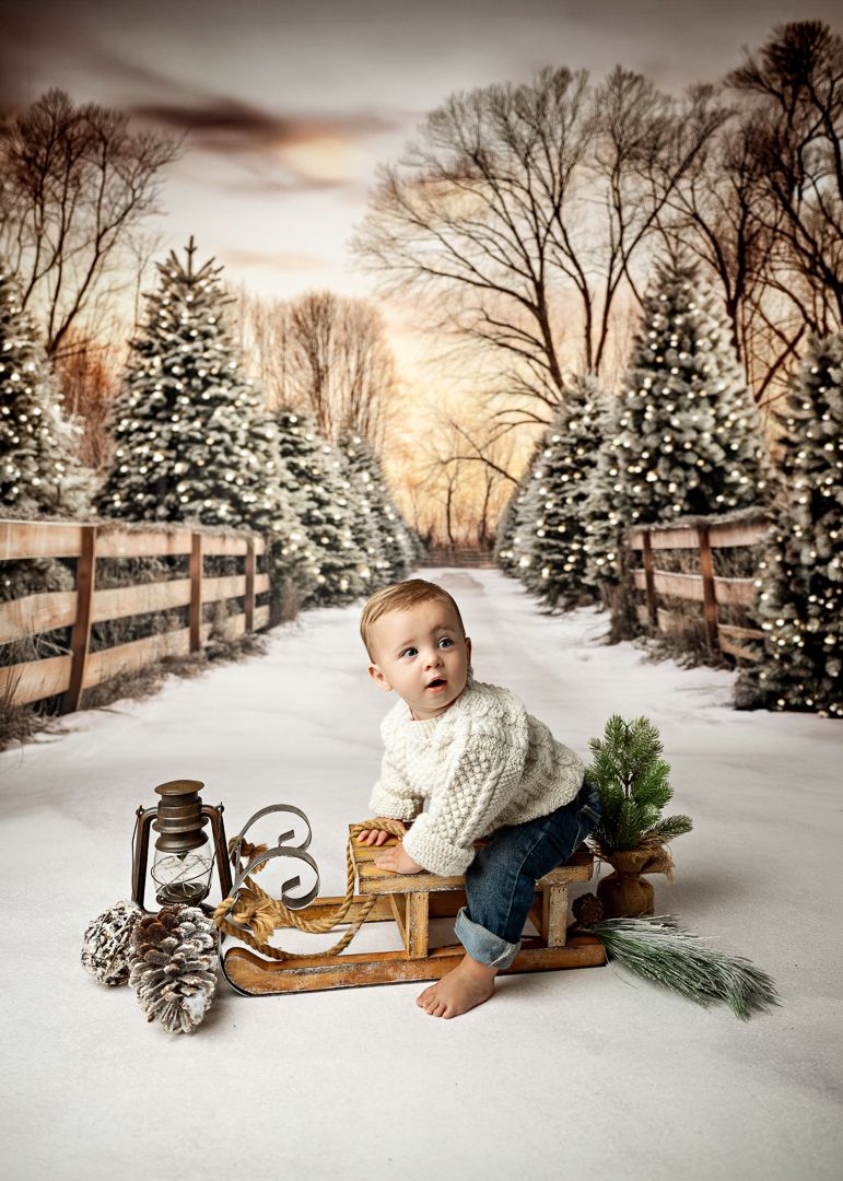 Baby in white sweater sits on vintage sled with lantern and pinecones on snowy Christmas backdrop.