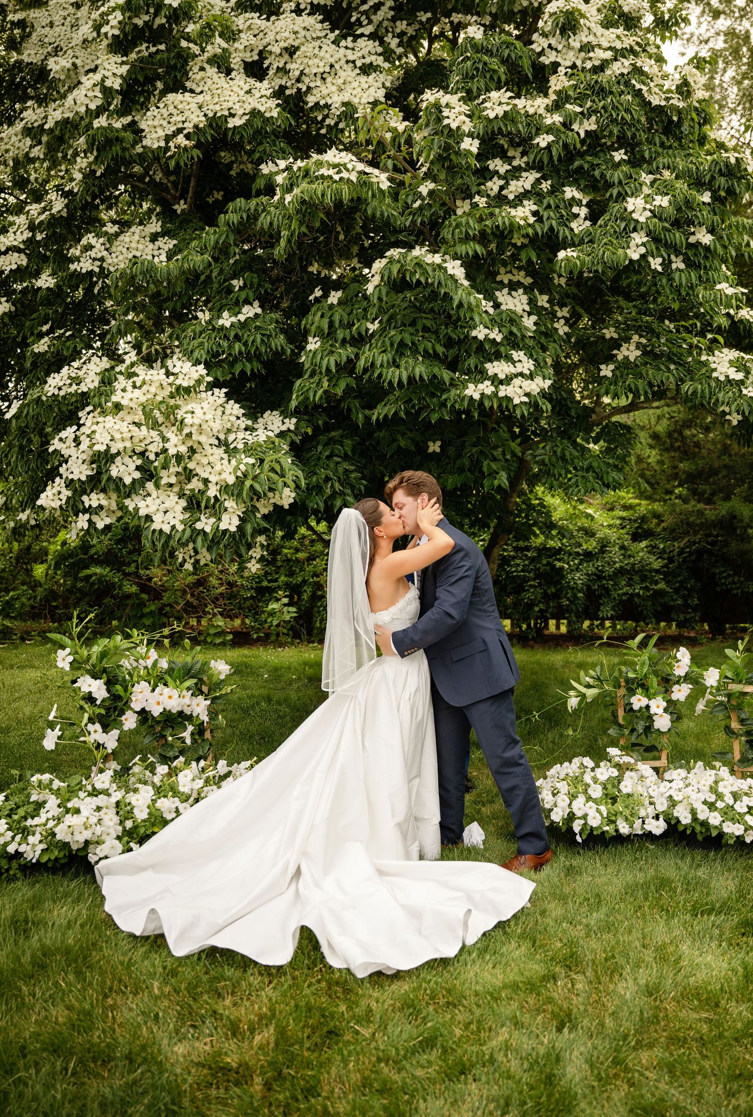 Bride and groom share a kiss outdoors surrounded by white flowers and lush green trees during their wedding ceremony.