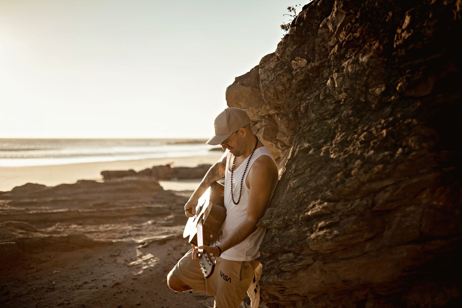 A person stands against a rocky cliff at sunset on a scenic beach, with golden light illuminating the coastal scene.