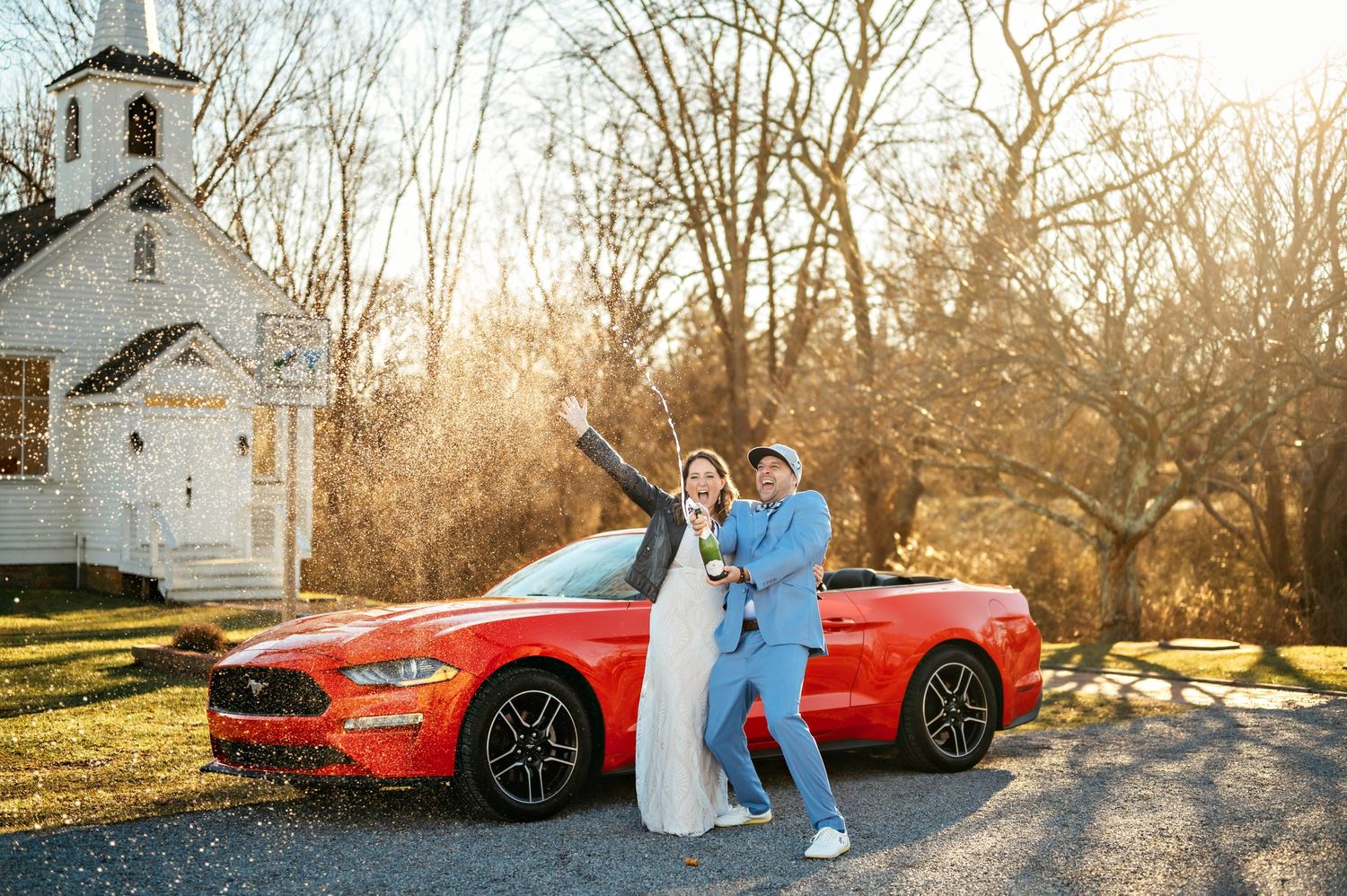 Couple celebrates with vintage white church and red Ford Mustang convertible in golden sunlight.