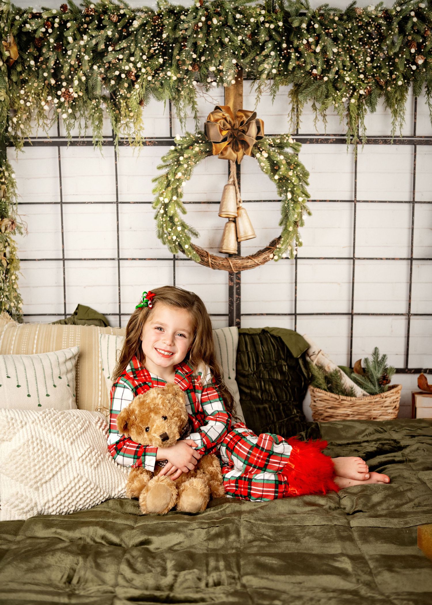 Holiday portrait scene with plaid pajamas and teddy bear against rustic backdrop with greenery and wreath decorations.