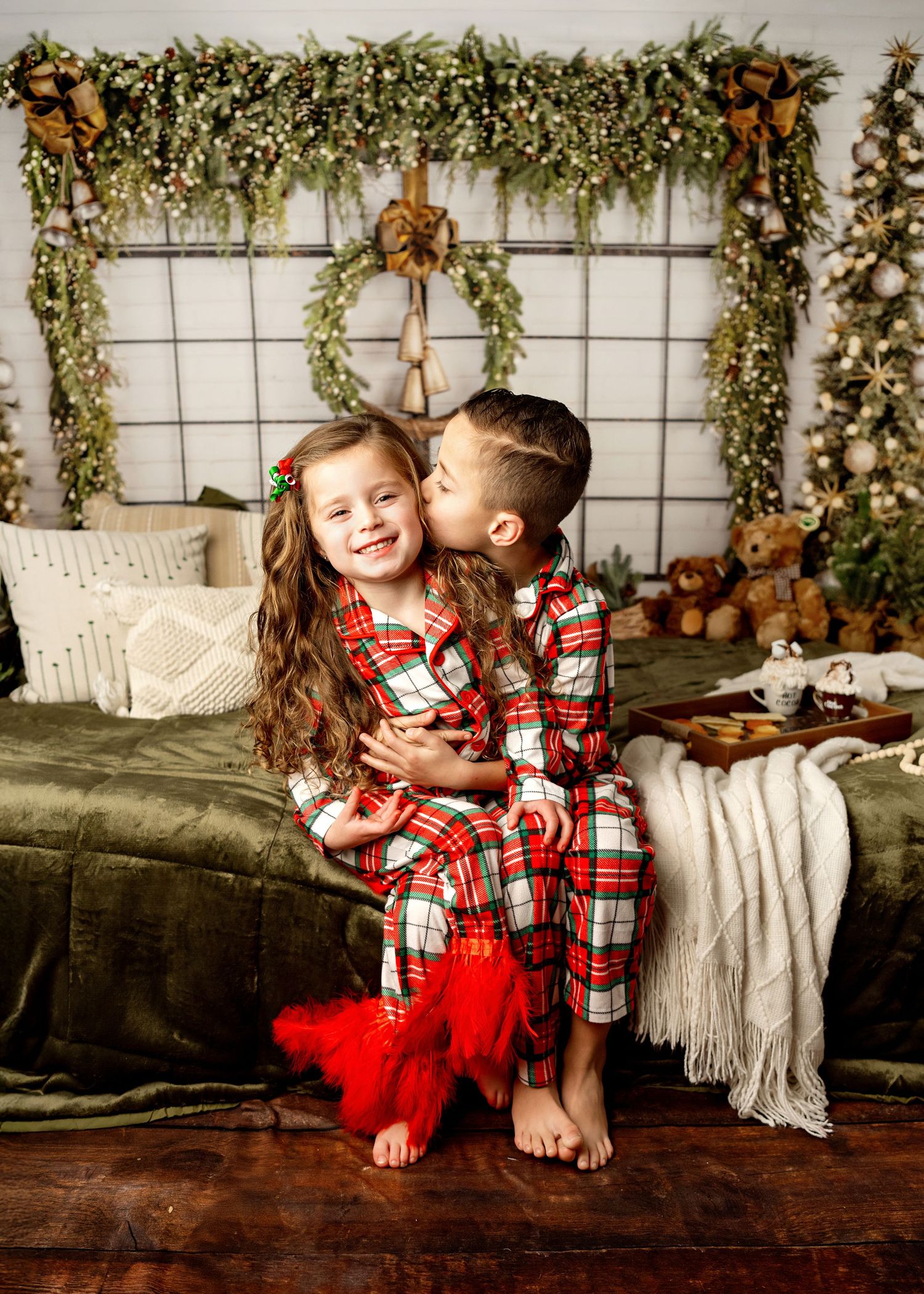 Two children in matching Christmas plaid pajamas share a sweet moment on a couch with festive holiday decor backdrop.