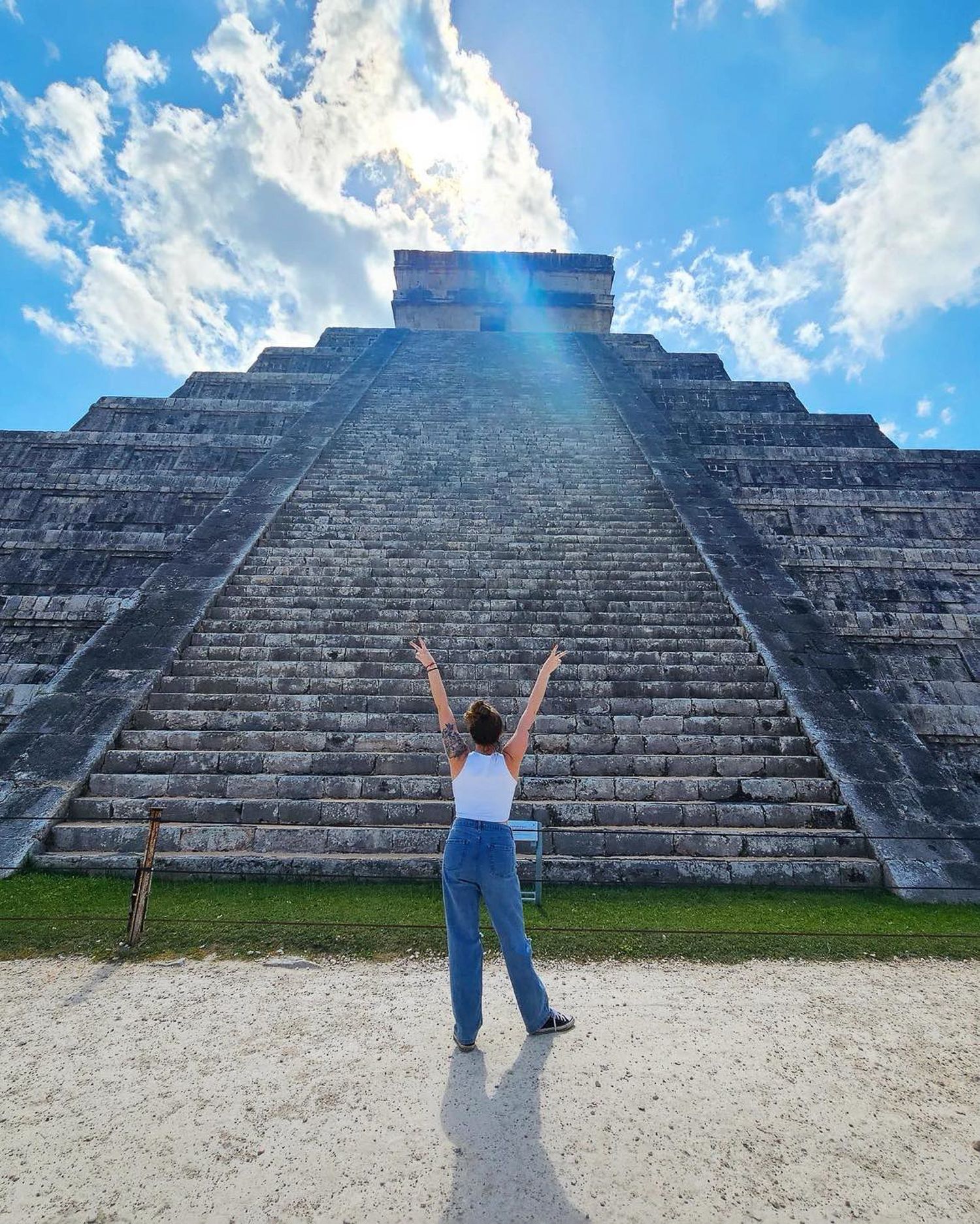 A visitor with raised arms stands before the majestic El Castillo pyramid at Chichen Itza against a bright blue sky.