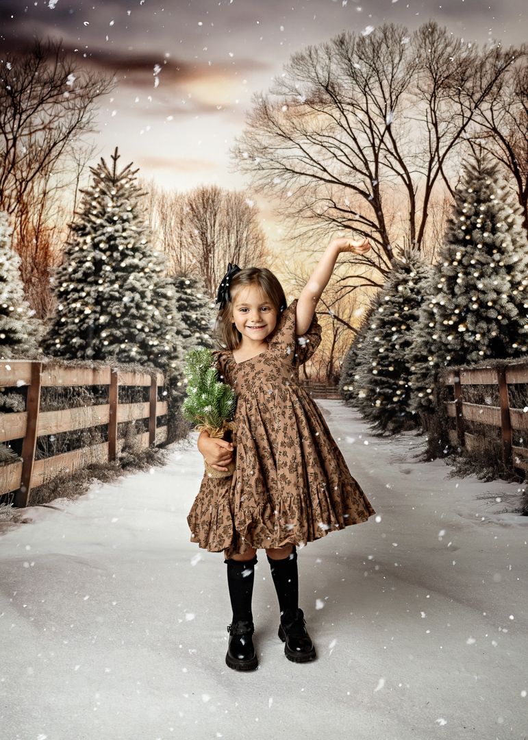 Young girl in a floral dress holds a small Christmas tree while snow falls at a festive tree farm.