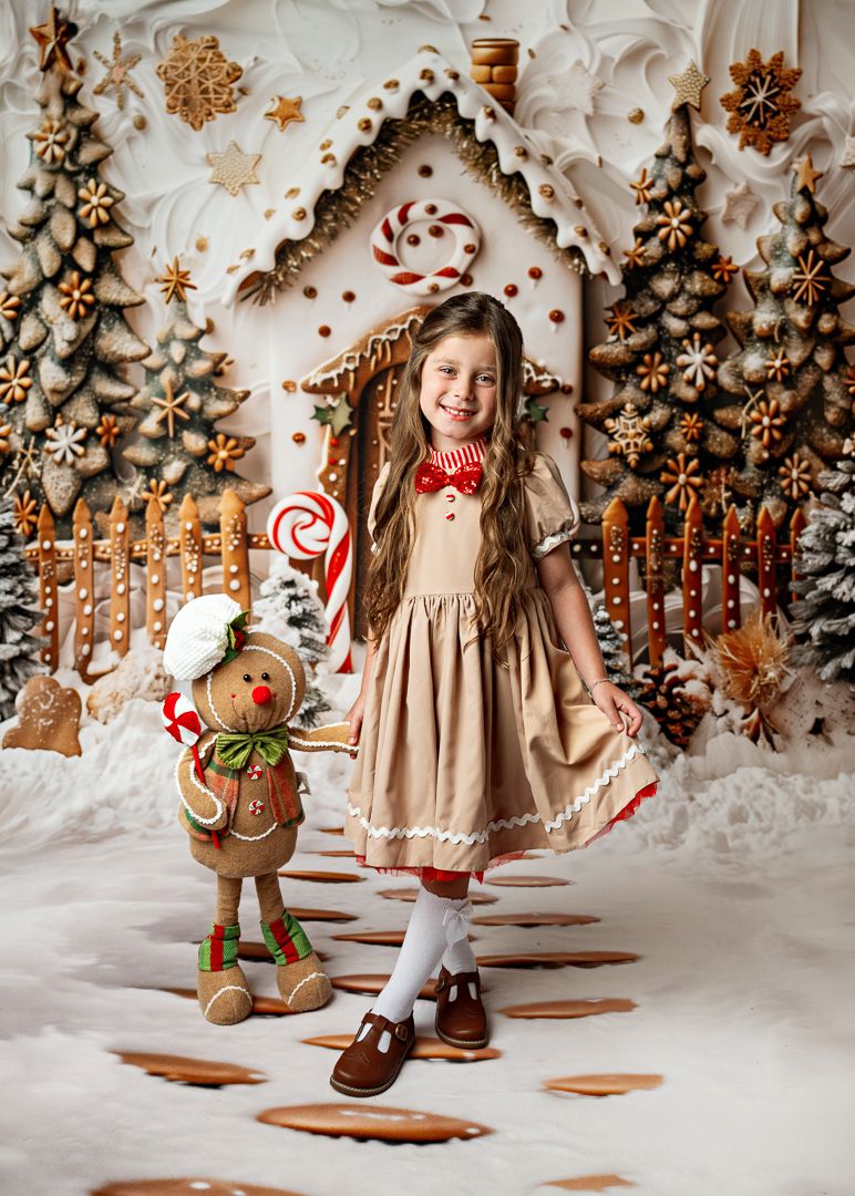 Young girl in a tan dress smiles beside a gingerbread man figure in a whimsical Christmas candy house setting.