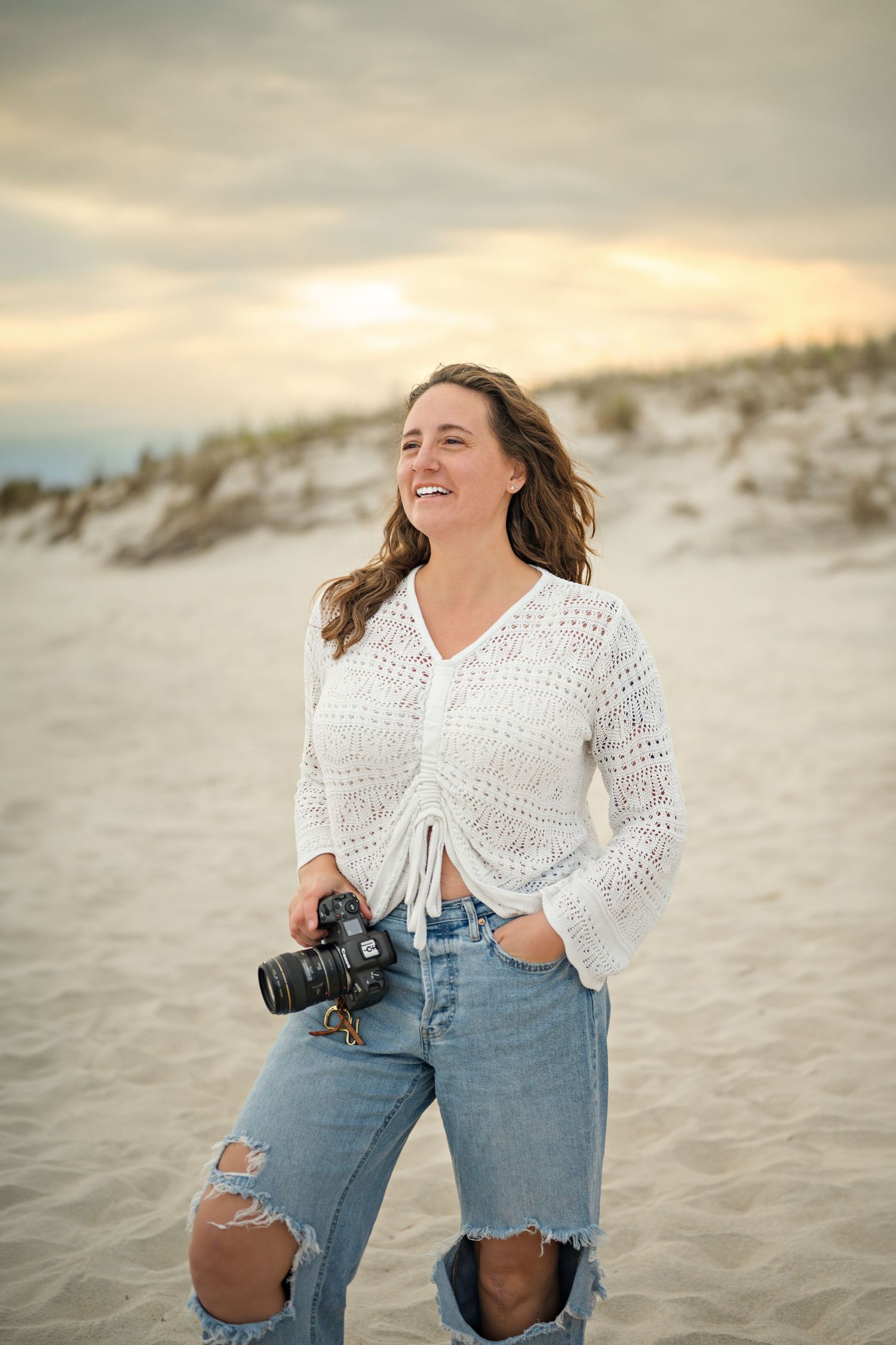 Woman in white crochet top and ripped jeans holding a camera smiling on a sandy beach at sunset.