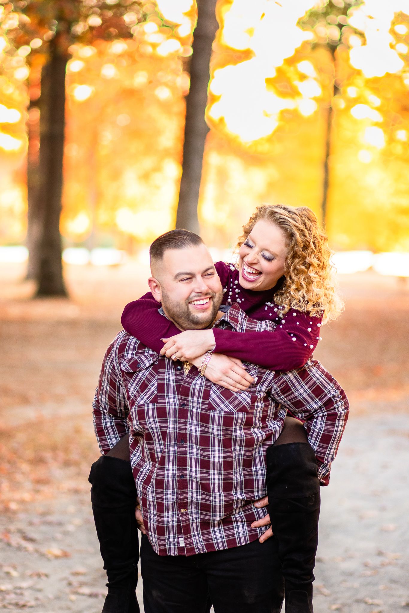 Playful autumn engagement photo with couple in matching burgundy amid golden sunlit trees in a park.