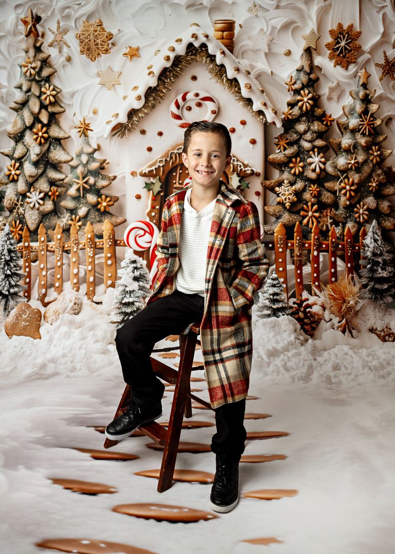 Smiling boy in plaid coat sits on stool before gingerbread house Christmas backdrop with candy canes and snow.