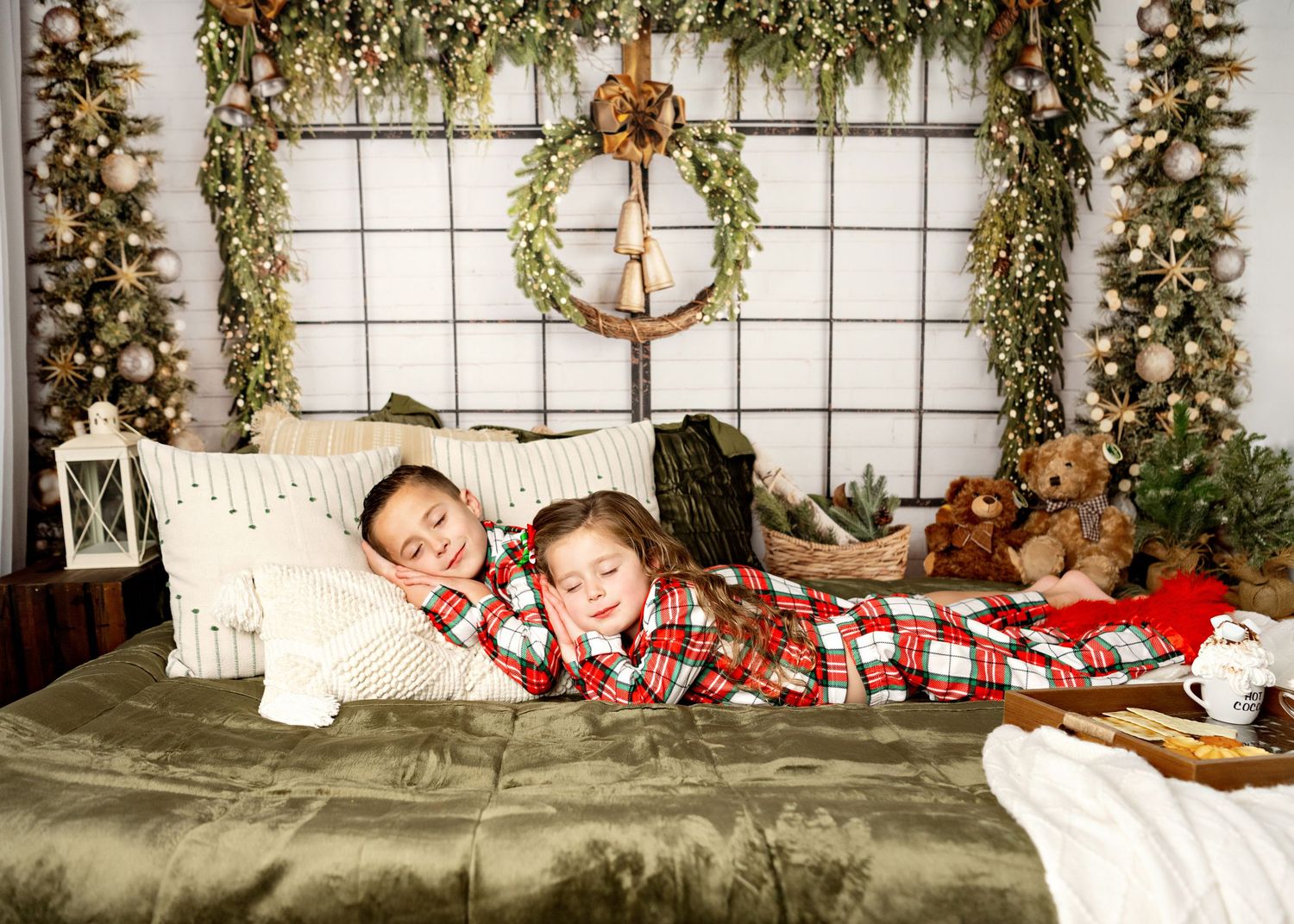 Two children in matching plaid pajamas lie on a bed decorated with holiday greenery and Christmas lights.