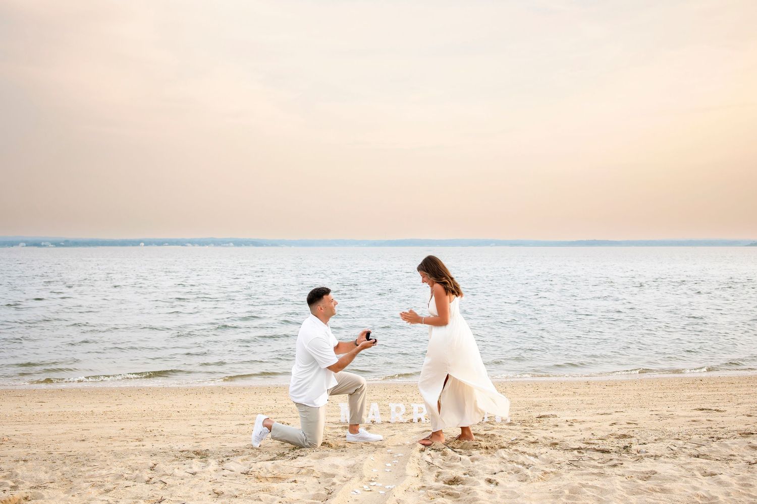 A couple in white clothing sits on a sandy beach during sunset while playing with their small dog.