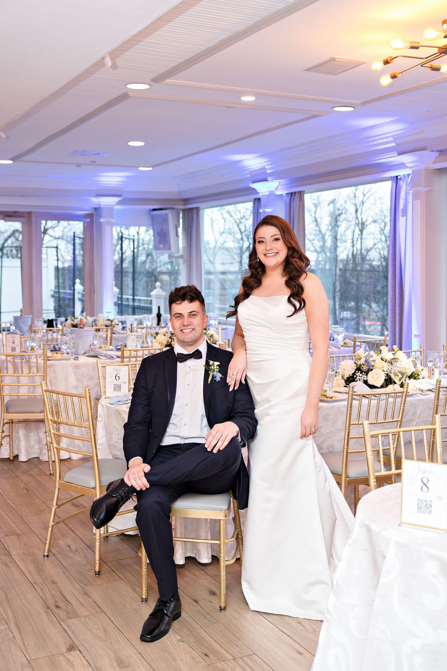 Bride and groom pose elegantly in a decorated wedding reception hall with gold chairs and floral centerpieces.