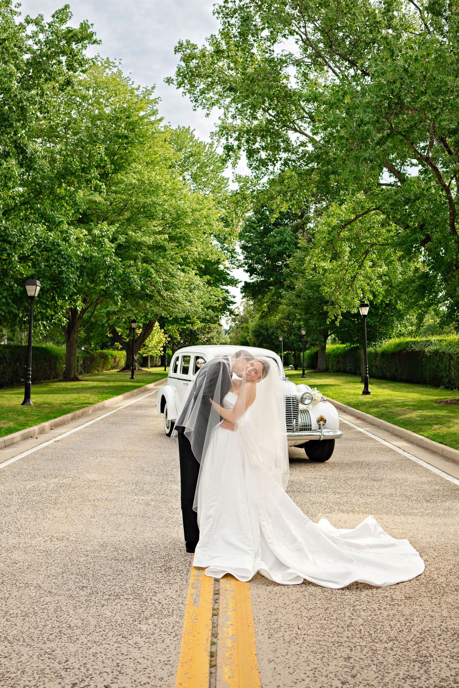 Bride and groom kissing on tree-lined road with vintage white classic car backdrop on their wedding day.
