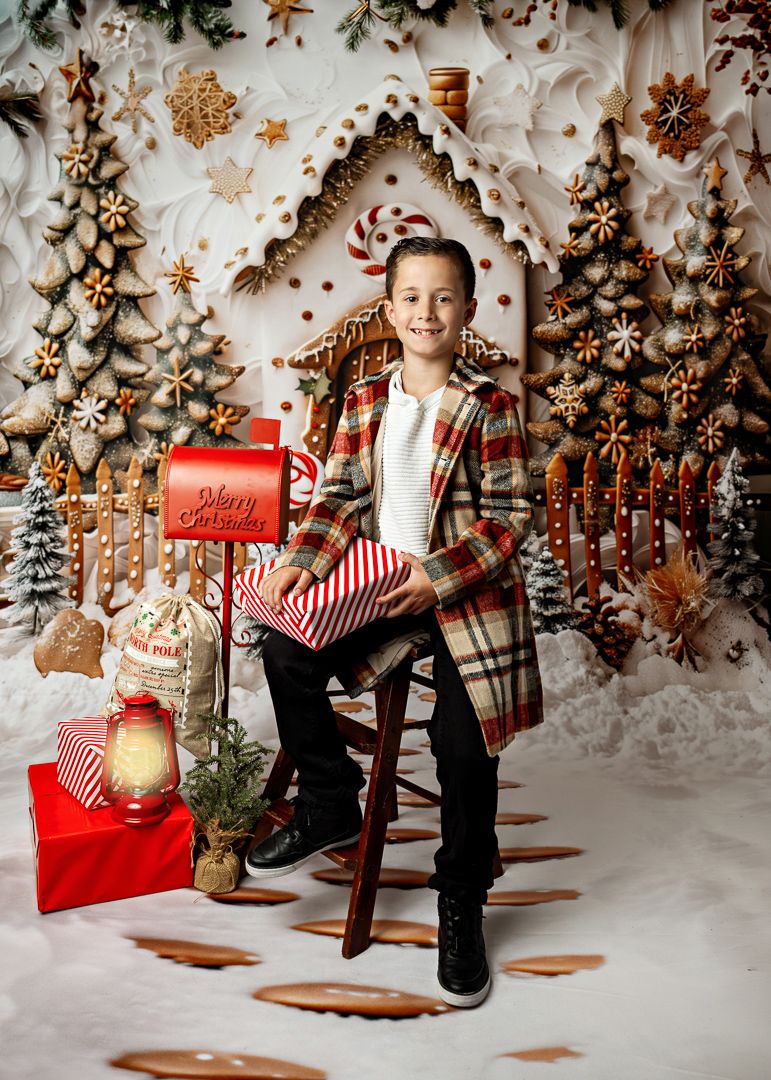 Smiling boy in plaid coat holds gift beside red mailbox with gingerbread house Christmas backdrop and holiday decor.