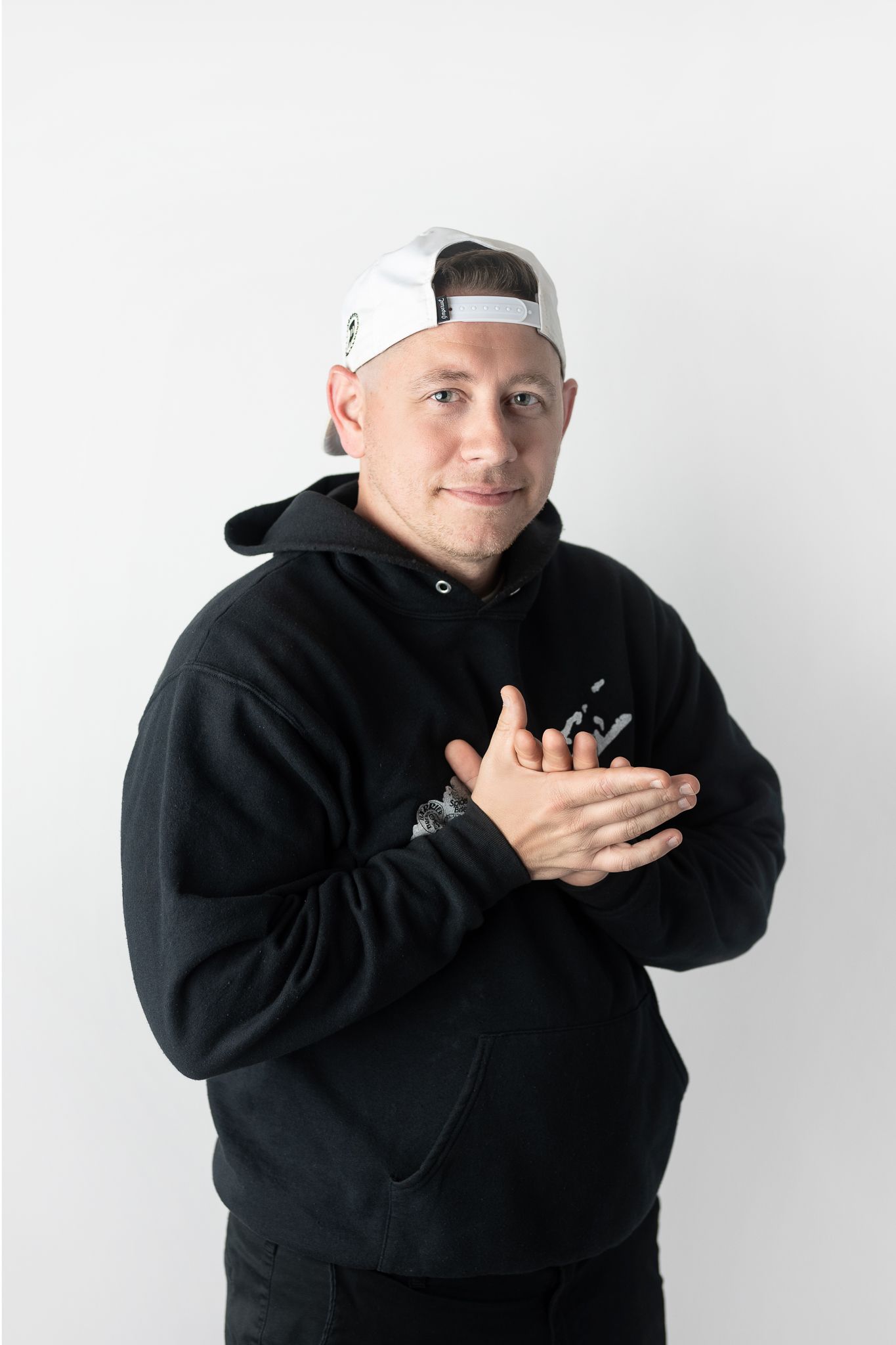 Person wearing a backwards white cap and black hoodie poses against white background with hands crossed over chest.