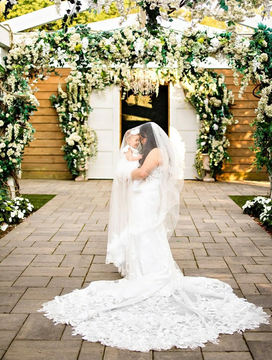 Bride in lace wedding gown and veil stands before floral arch with white flowers at elegant outdoor venue.