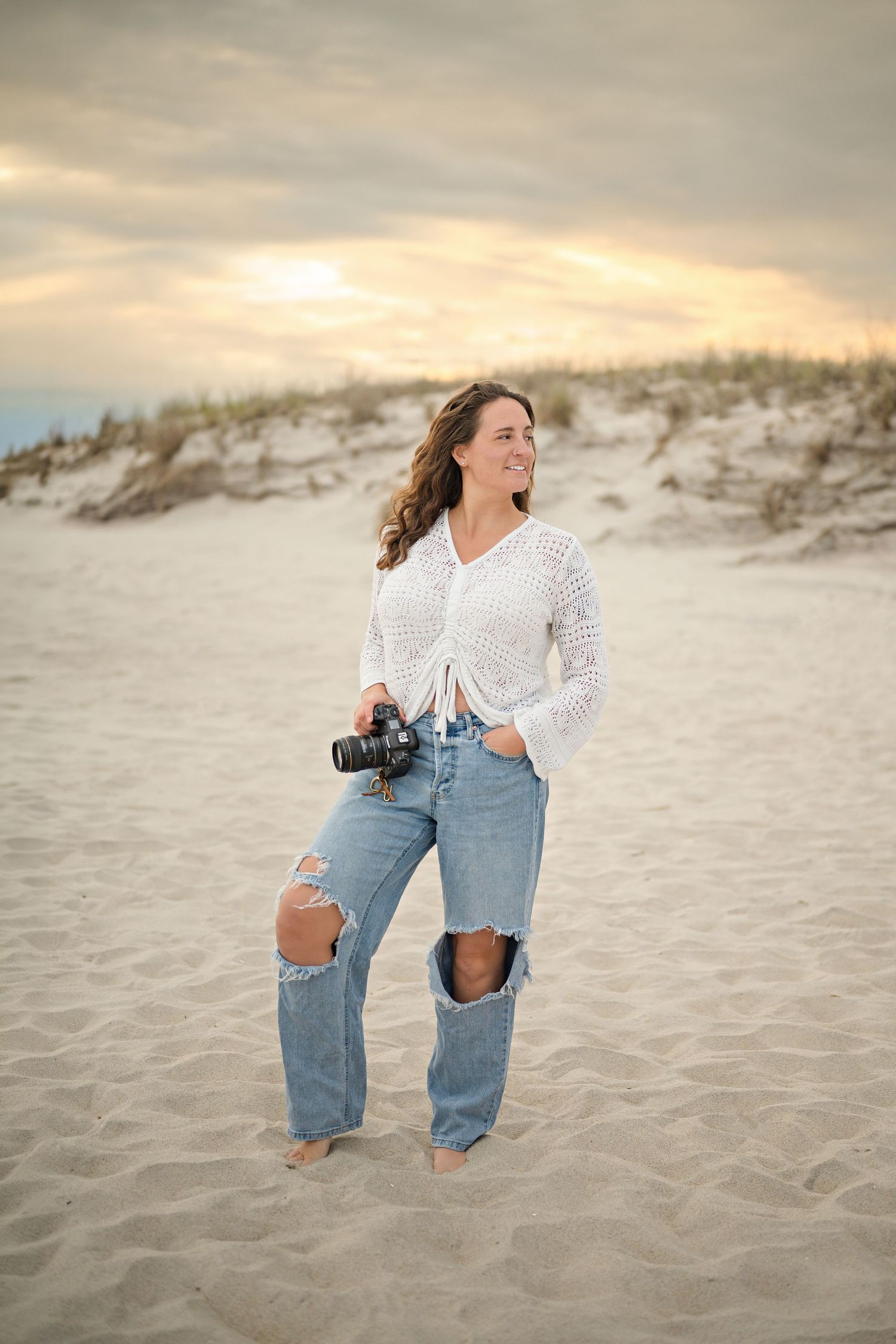 Woman photographer holding camera on sandy beach at sunset, wearing white top and ripped jeans, smiling.