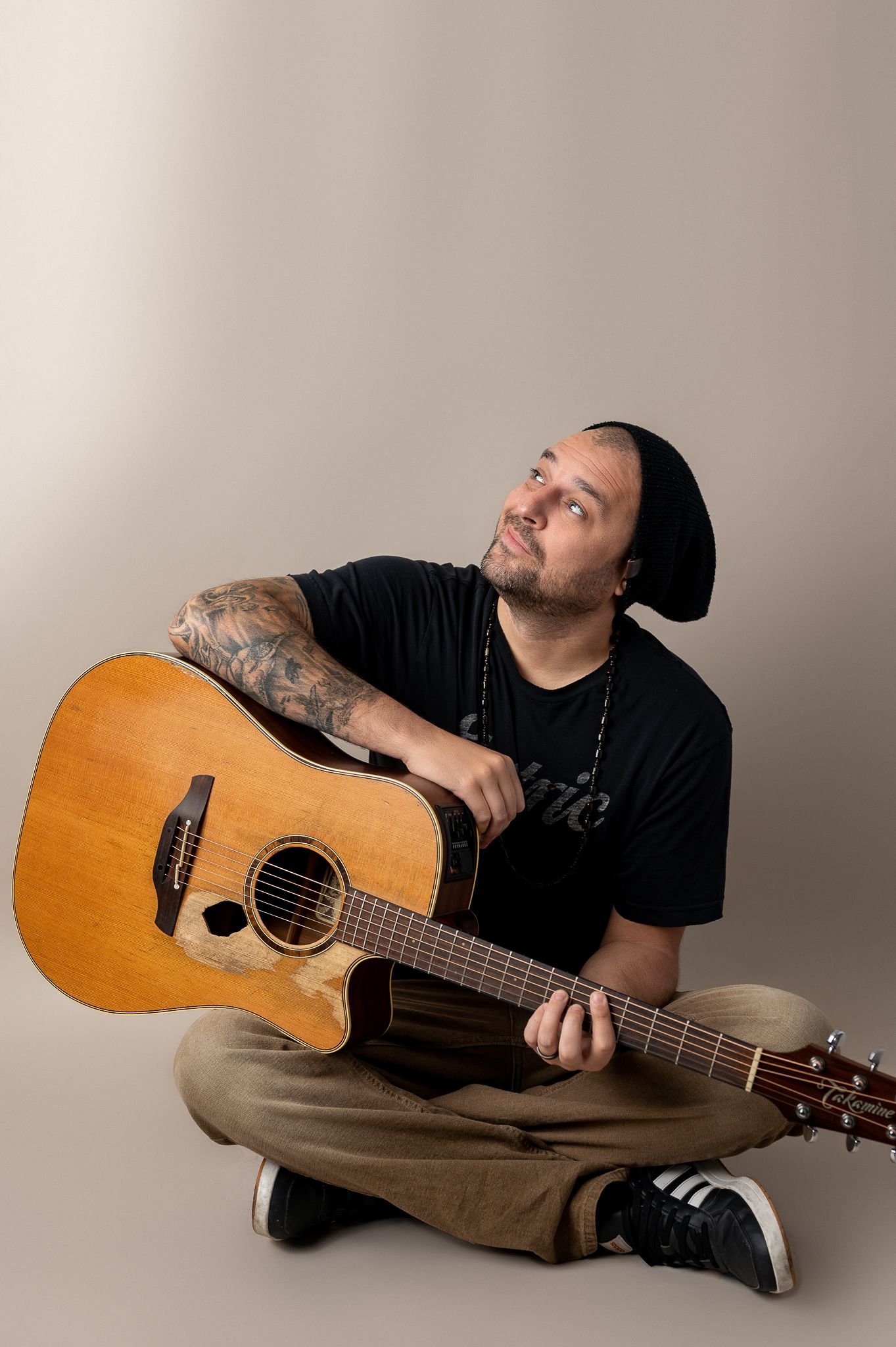 Musician in black t-shirt and beanie sits cross-legged with acoustic guitar against neutral backdrop.