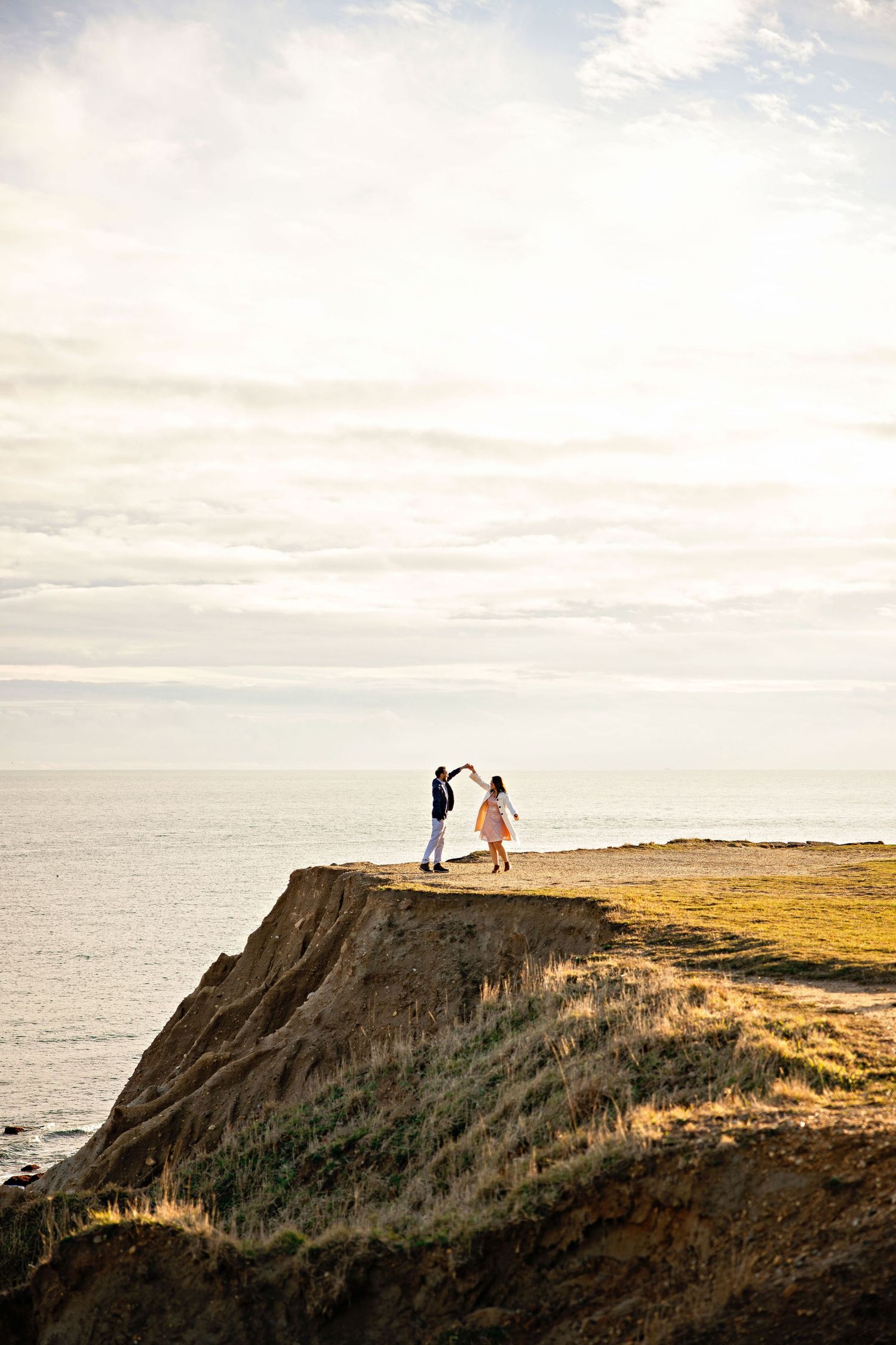Two people stand together on a dramatic coastal cliff overlooking the ocean at sunset with soft clouds in the sky.