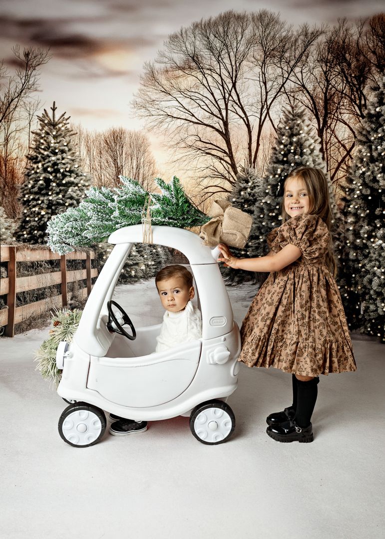 Two children pose in a snowy Christmas tree farm scene, one in a toy car with a tree on top.