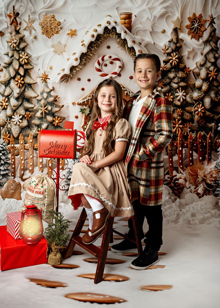 Two children pose in front of a festive gingerbread Christmas backdrop with a red Merry Christmas mailbox.