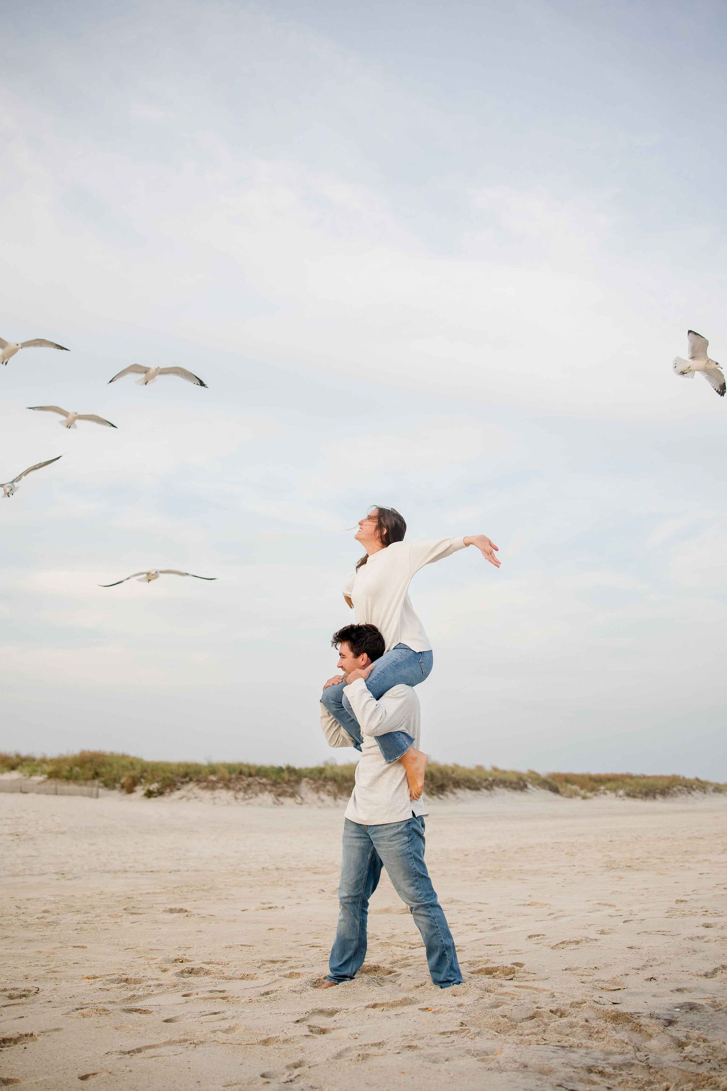 Person in jeans plays with seagulls on a sandy beach as birds soar overhead against a cloudy sky.