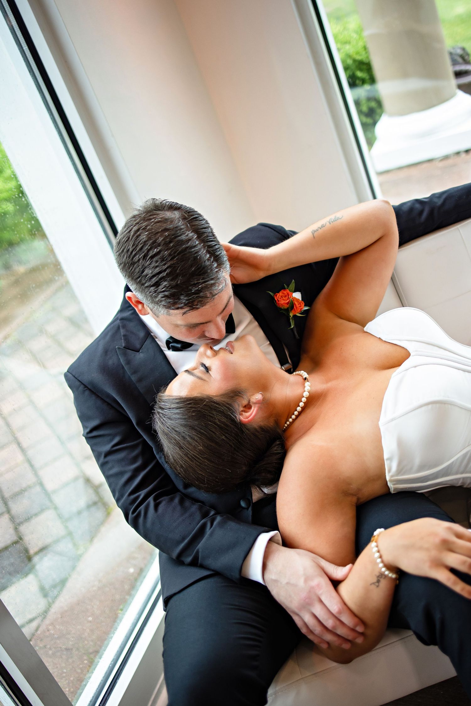 A bride and groom share a romantic kiss while reclining on a couch near large windows at their wedding reception.