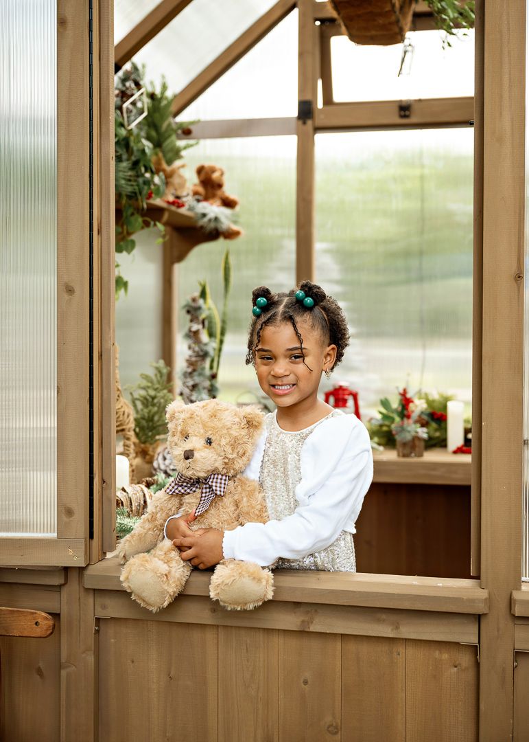 Happy young girl holding a teddy bear in the window of a Christmas-decorated wooden greenhouse.