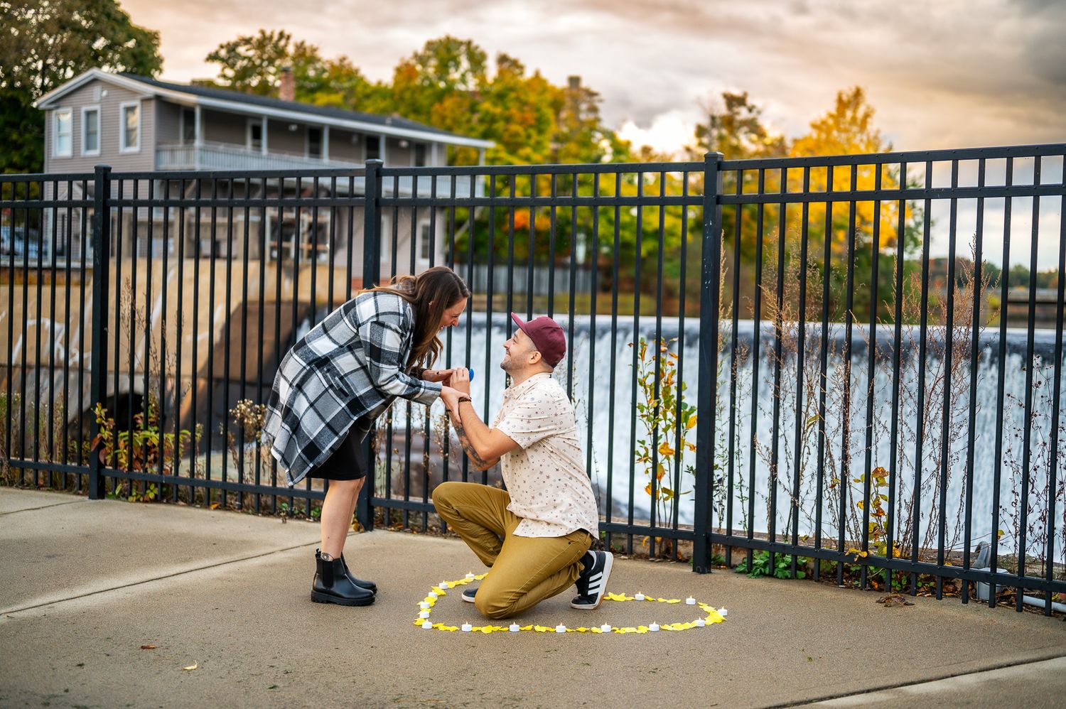 Two people share a sweet moment at sunset by a lake with a metal railing, surrounded by autumn trees in golden light.