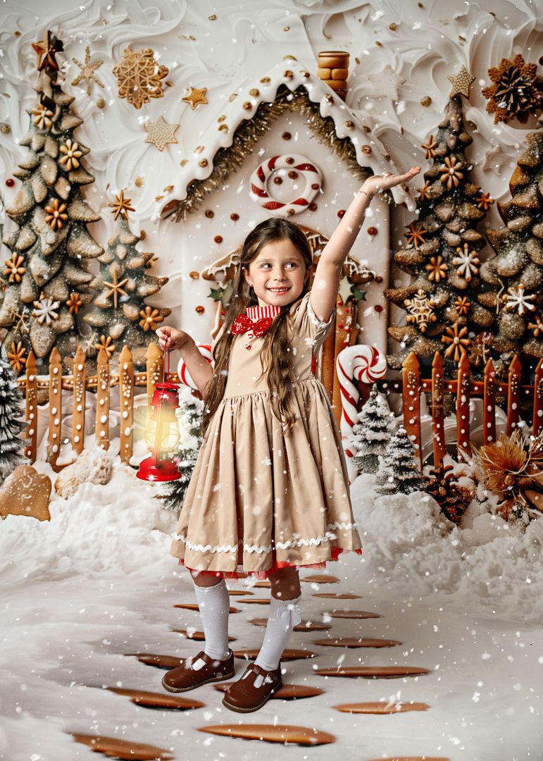 Young girl in beige dress holds lantern, arm raised, posing in front of festive gingerbread house Christmas backdrop.